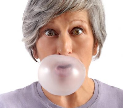 Adult Woman with Short Gray Hair Blowing Chewing Gum with Eyes Wide Open Looking at Camera. Isolated on White Background.