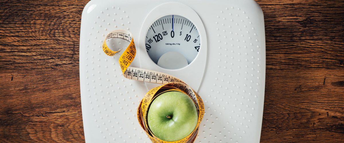 Green apple wrapped in a tape measure on a white scale, wooden surface on background, dieting and weight loss concept