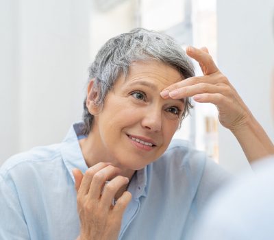 Mature woman looking her face and wrinkles in the bathroom mirror. Senior woman applying cosmetic lotion on skin between eyebrows while looking at mirror. Beautiful lady looking her face in the early morning.