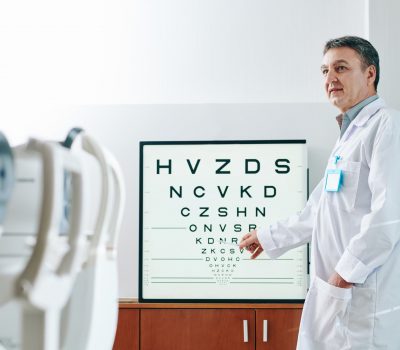 Mature ophthalmologist pointing at letters on test chart when checking eyesight of patient in clinic