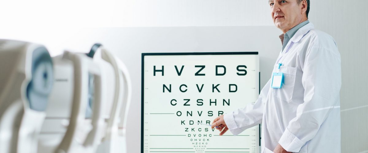 Mature ophthalmologist pointing at letters on test chart when checking eyesight of patient in clinic