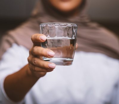 Unrecognizable Muslim Woman Offering Glass Of Water To Camera Standing In Modern Kitchen At Home, Wearing Hijab. Healthy Hydration, Drink More Water And Stay Hydrated. Selective Focus, Cropped